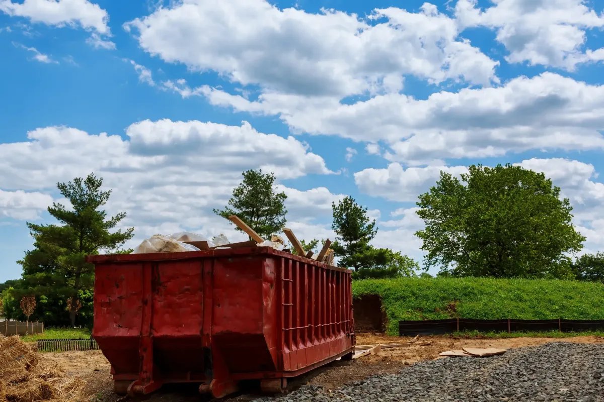 Red dumpster filled with debris under a blue sky with clouds, surrounded by trees and construction materials, representing efficient waste removal for home projects.