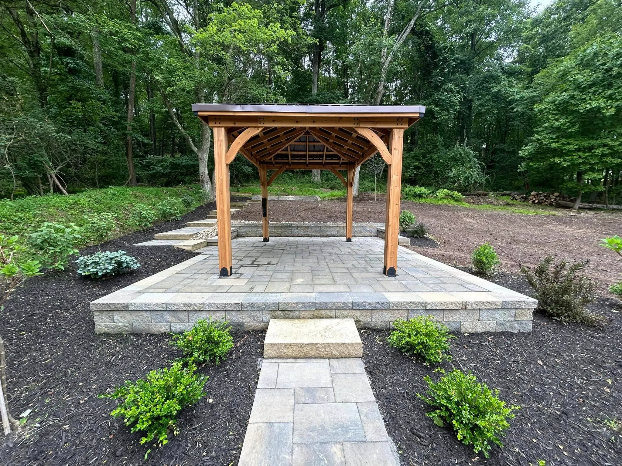 Wooden gazebo on stone patio surrounded by landscaped garden, featuring greenery and mulch, ideal for outdoor gatherings and enhancing property aesthetics.