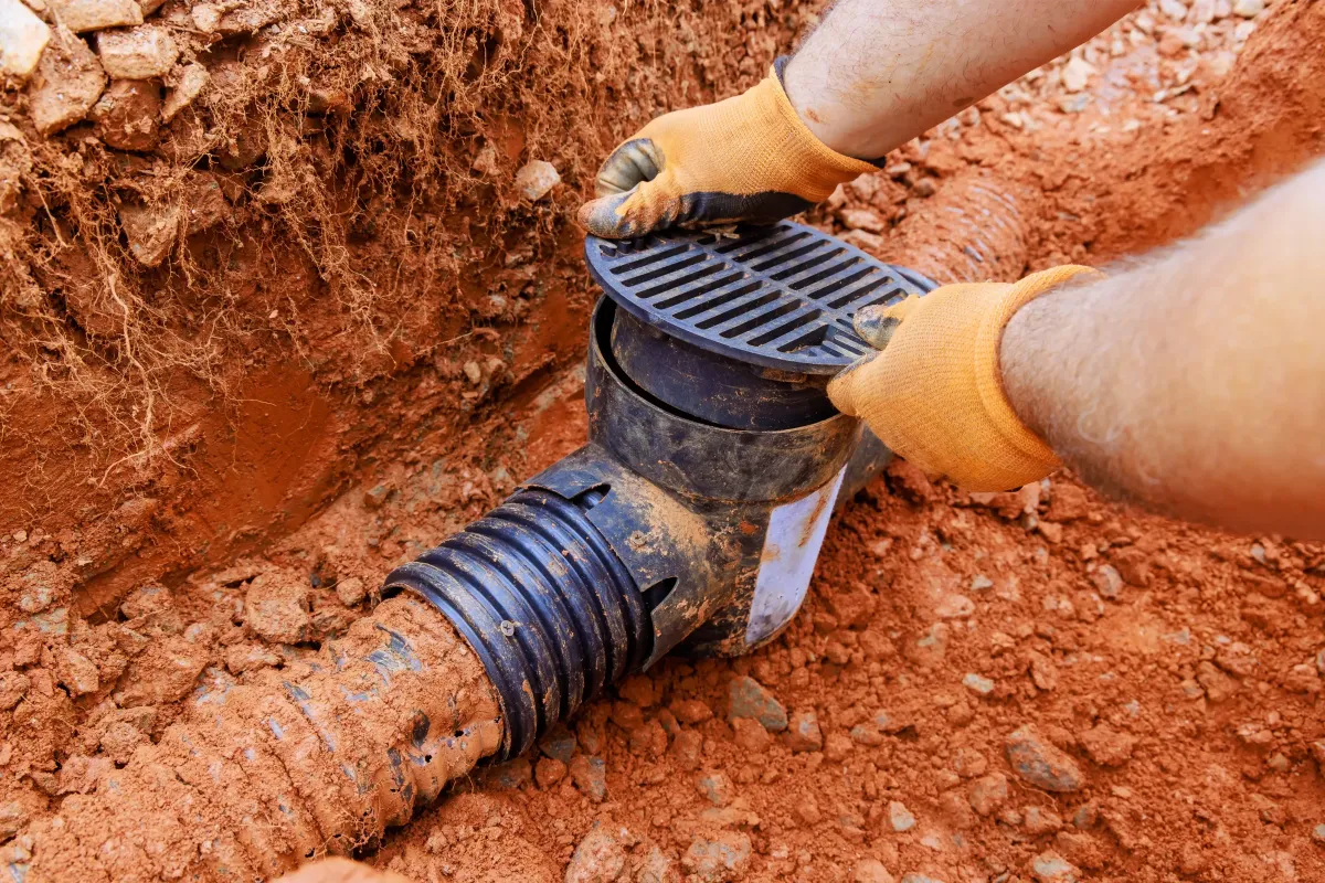Person wearing orange gloves installing a drainage system in red soil, emphasizing water management solutions for landscaping.