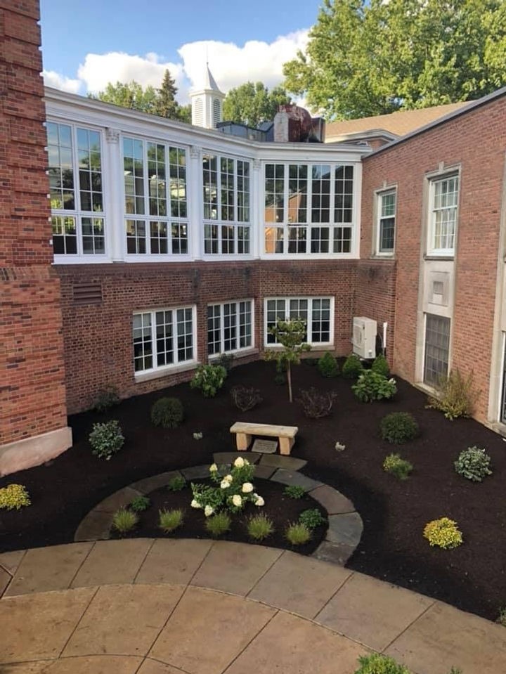 Landscaped courtyard featuring a stone bench, circular flower bed with white flowers, surrounded by lush greenery and brick buildings, showcasing a serene outdoor space for landscaping projects.