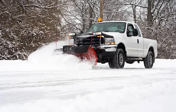 White pickup truck with snow plow clearing snow-covered road in winter landscape, illustrating snow removal services.