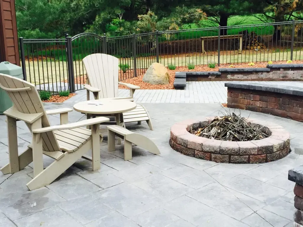 Patio area featuring wooden Adirondack chairs and a round table surrounding a stone fire pit, set in a landscaped outdoor space with greenery and decorative stones.