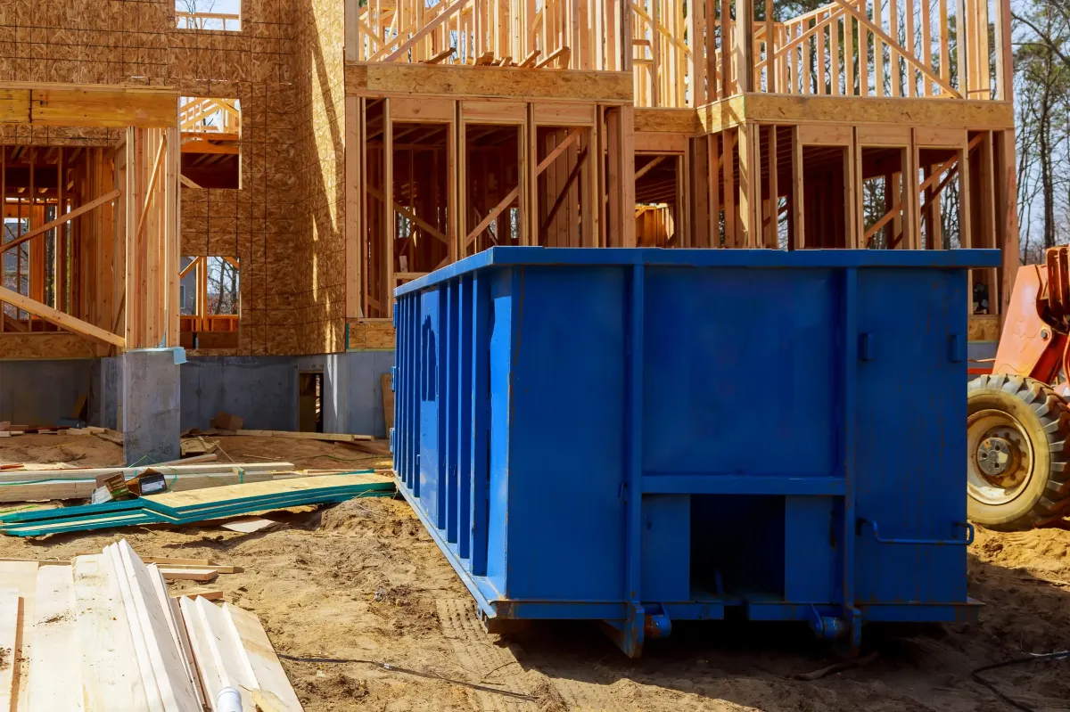 Blue dumpster on construction site surrounded by wooden framing and construction materials, illustrating efficient waste disposal for landscaping and construction projects.